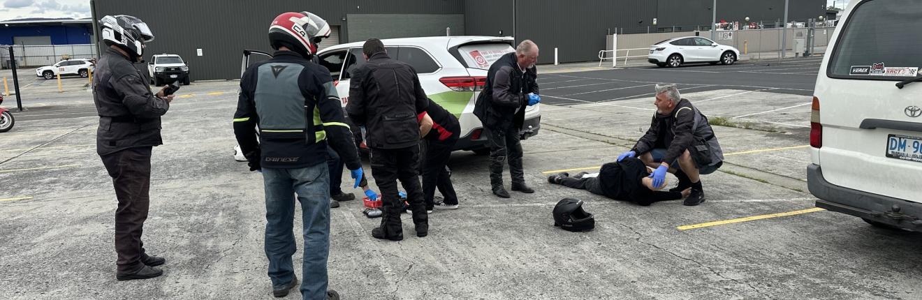  This photo shows a group of motorcyclists participating in a first aid training session in a car park. Several individuals wearing motorcycle gear and helmets are standing around, while others are actively involved in assisting a person lying on the ground. 