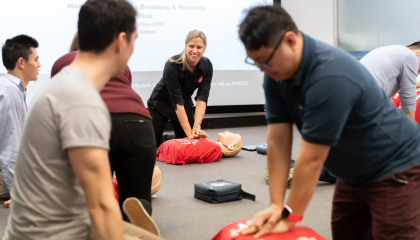 A trainer demonstrating CPR on manikins in the background, with students practicing in the foreground. 
