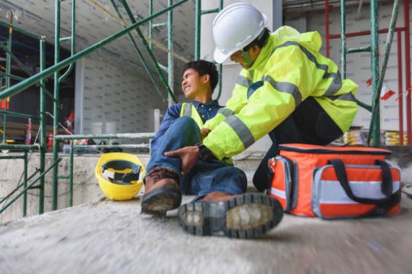 Two people on a work site, one is lying down in pain, the other is administering first aid